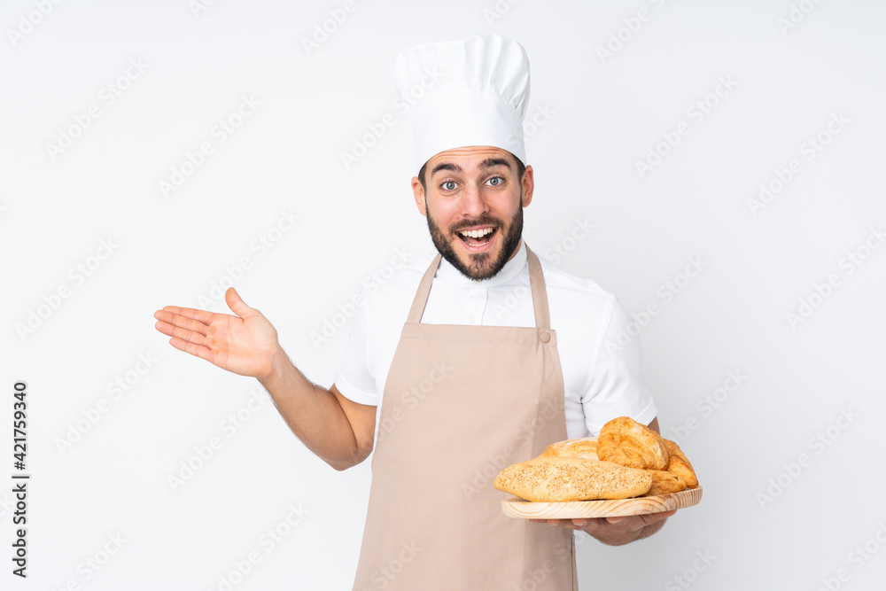 Male baker holding a table with several breads isolated on white background extending hands to the side for inviting to come Male baker holding a table with several breads isolated on white background extending hands to the side for inviting to come