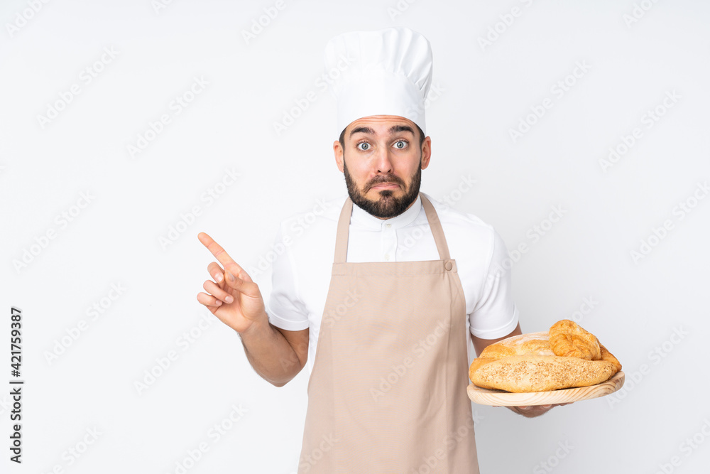 Male baker holding a table with several breads isolated on white background pointing to the laterals having doubts Male baker holding a table with several breads isolated on white background pointing to the laterals having doubts