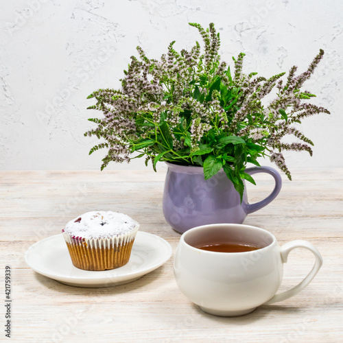 White mug of tea on a background of muffin and mint flowers on a white wooden table. Tea party with a cupcake.