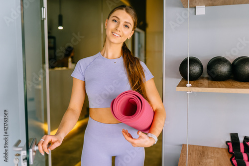 Portrait of excited female yoga teacher posing with pink fitness mat, happy toned fitness instructor or coach in lilac sportswear smiling making picture before training in fitnesss studio. photo