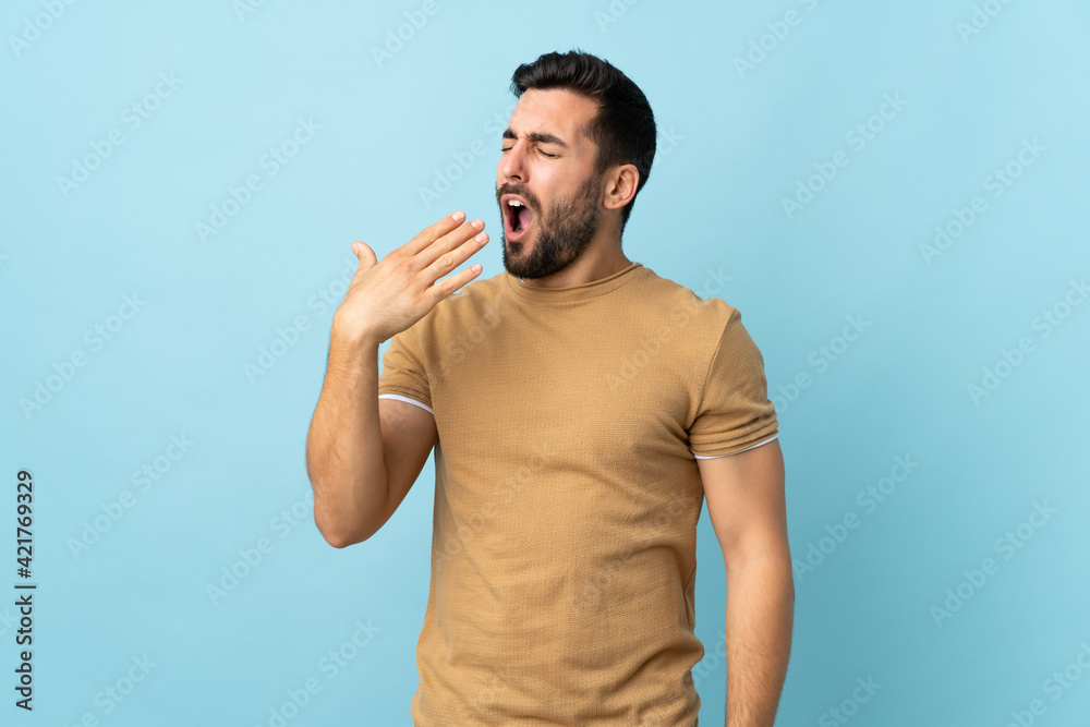 Young handsome man with beard over isolated background yawning and covering wide open mouth with hand Young handsome man with beard over isolated background yawning and covering wide open mouth with hand