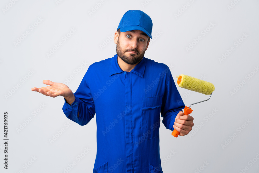Painter man holding a paint roller isolated on white background having doubts while raising hands