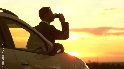 A businessman in a fake suit drinks coffee at the evening sunset in the sky standing next to the car  freedom of travel behind the wheel  concentrate and think about the future outdoors  a man at dawn