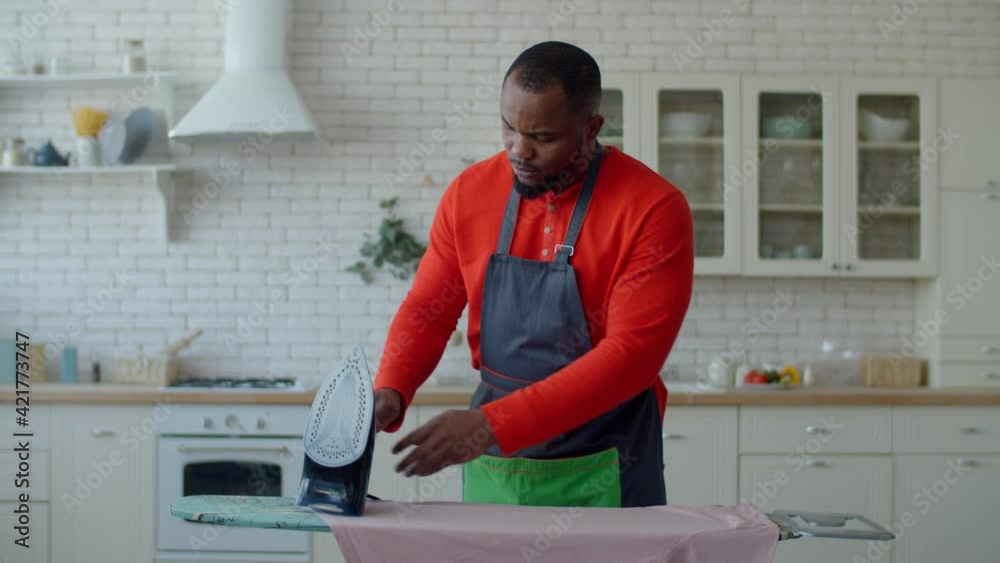 Concentrated multitasking attractive black male in apron making housekeeping duties, ironing shirt with electric iron and answering phone call while doing household chores in domestic room.