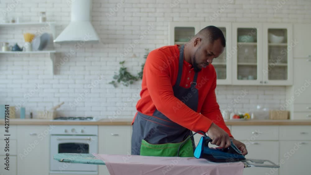 Serious handsome african american single man in apron doing household chores, ironing clothes with electric iron on ironing board while making housekeeping duties in home interior during weekend. Serious handsome african american single man in apron doing household chores, ironing clothes with electric iron on ironing board while making housekeeping duties in home interior during weekend.