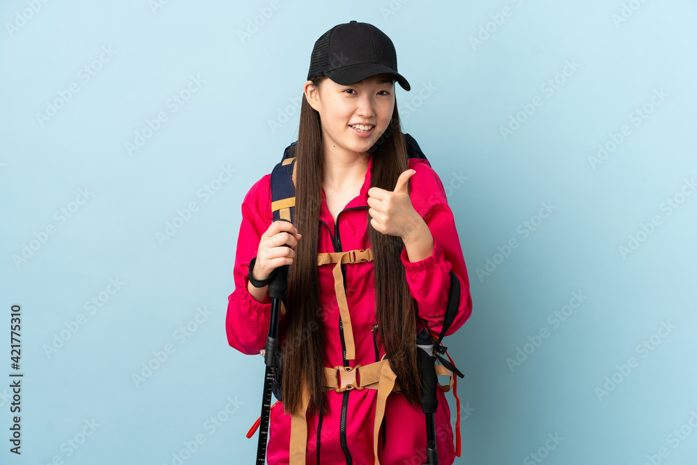 Young Chinese girl with backpack and trekking poles over isolated blue background giving a thumbs up gesture