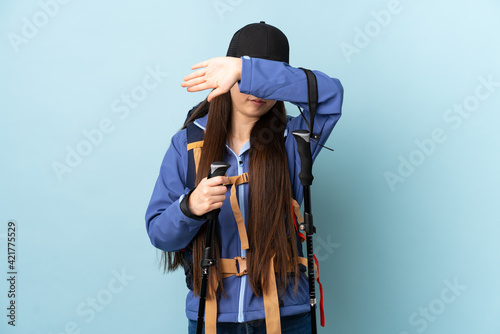 Young Chinese girl with backpack and trekking poles over isolated blue background covering eyes by hands