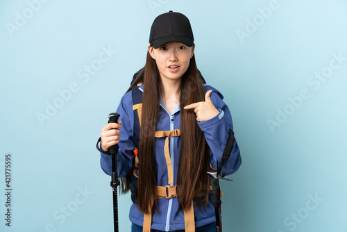 Young Chinese girl with backpack and trekking poles over isolated blue background with surprise facial expression