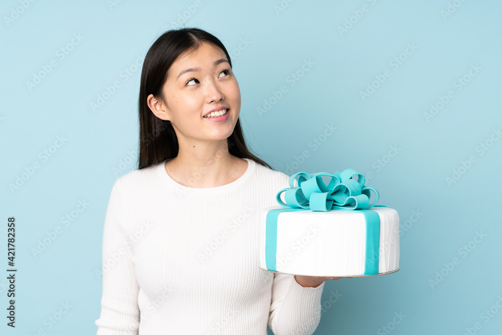 Pastry asian chef holding a big cake isolated on blue background looking up while smiling Pastry asian chef holding a big cake isolated on blue background looking up while smiling