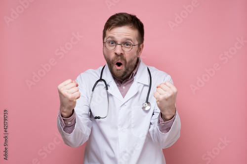 Satisfied male doctor in a white coat  with a stethoscope around neck  makes a gesture with hands and shouts joyfully. Pink background.