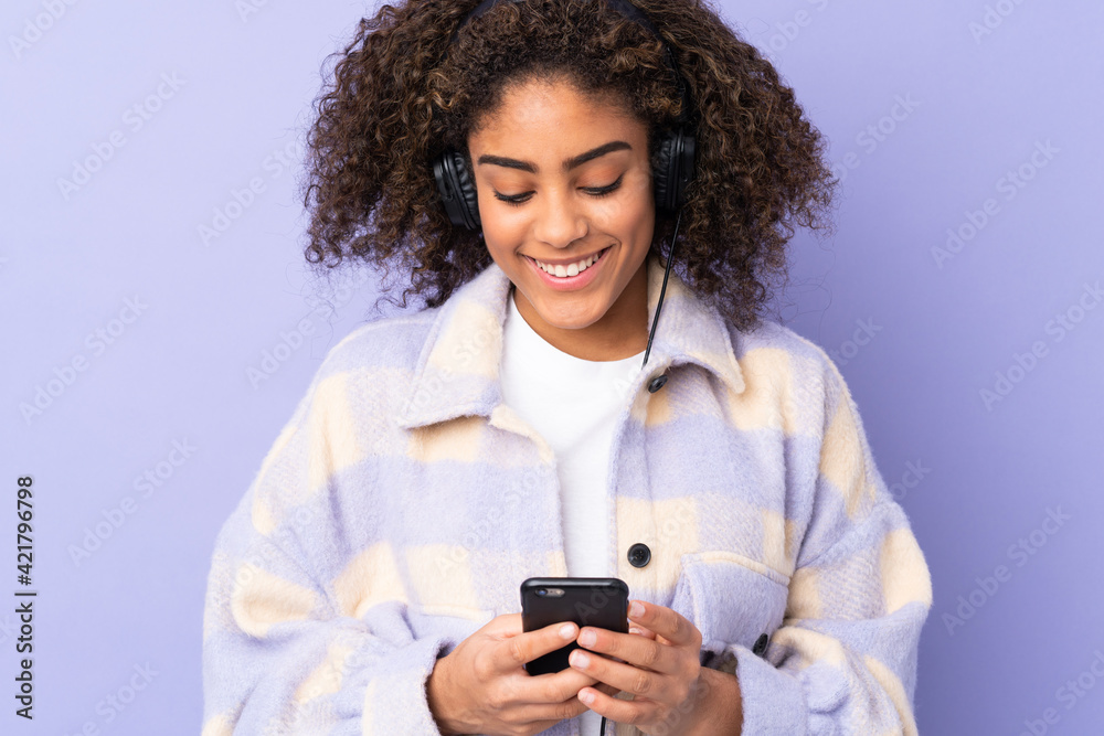 Young African American woman isolated on purple background listening music and looking to mobile Young African American woman isolated on purple background listening music and looking to mobile