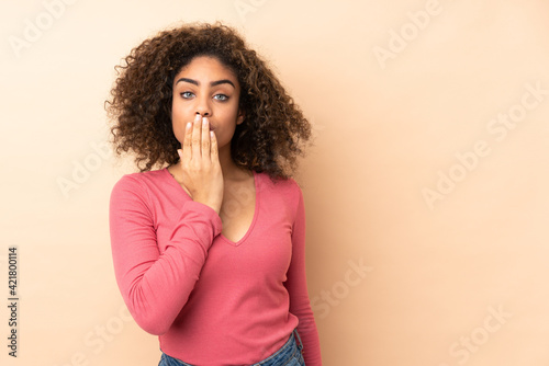 Young African American woman isolated on beige background covering mouth with hand