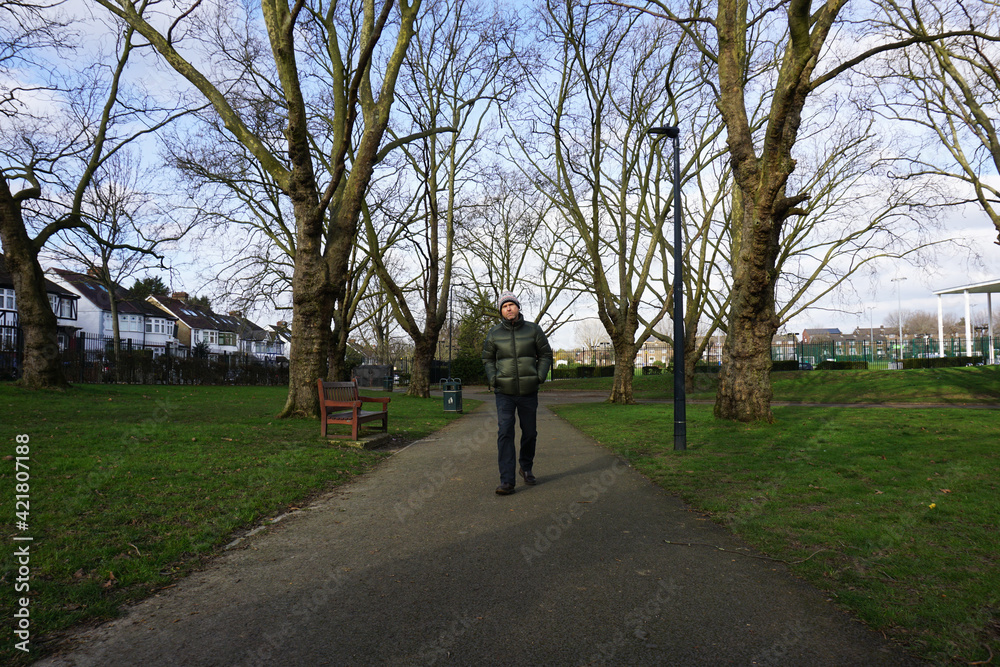 Portrait full body of Caucasian man wearing green jacket, bobble hat with white pom pom walking on pathway in the park, leafless tree under blue sky Portrait full body of Caucasian man wearing green jacket, bobble hat with white pom pom walking on pathway in the park, leafless tree under blue sky