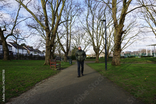 Portrait full body of Caucasian man wearing green jacket, bobble hat with white pom pom walking on pathway in the park, leafless tree under blue sky