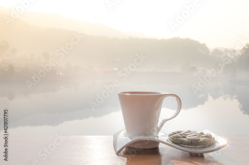 Coffee cup with nut cookie on the table with blurry nature village and lake background and morning  light flare. A good starting in the early morning to get ready for a day. Free copy space.