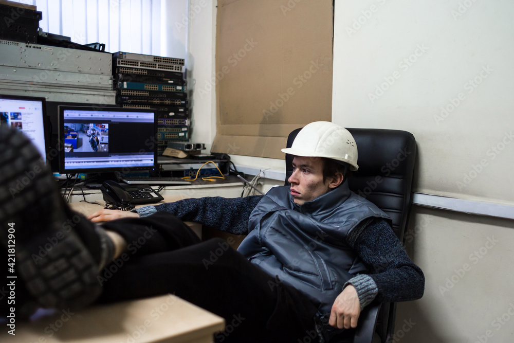 An engineer in a white helmet put his feet on the table at his workplace. Young worker is resting while sitting in a chair in front of monitors.