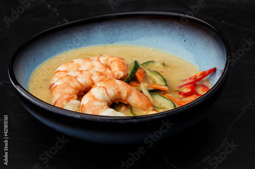 Asian soup with shrimps and vegetables: zucchini, red pepper and carrot. Dish isolated in a blue bowl, close-up on a black marble background. Asian cuisine.