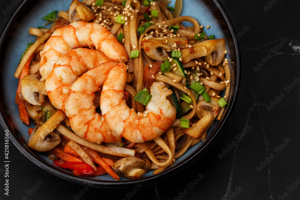Udon noodles with shrimps and vegetables: zucchini, red pepper, mushrooms, carrot, spring onion and sesame seeds. Dish isolated in a blue bowl, close-up on a black marble background. Asian cuisine. Udon noodles with shrimps and vegetables: zucchini, red pepper, mushrooms, carrot, spring onion and sesame seeds. Dish isolated in a blue bowl, close-up on a black marble background. Asian cuisine.