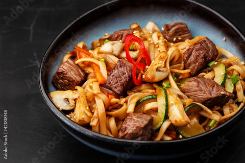 Udon noodles with teriyaki beef and vegetables: zucchini, red pepper, mushrooms, carrot, onion and sesame seeds. Dish isolated in a blue bowl, close-up on a black marble background. Asian cuisine.