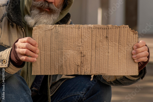Senior man homeless beggar with a gray beard in a shabby clothes with a .carboard sign sitting outdoors in city. Mock up