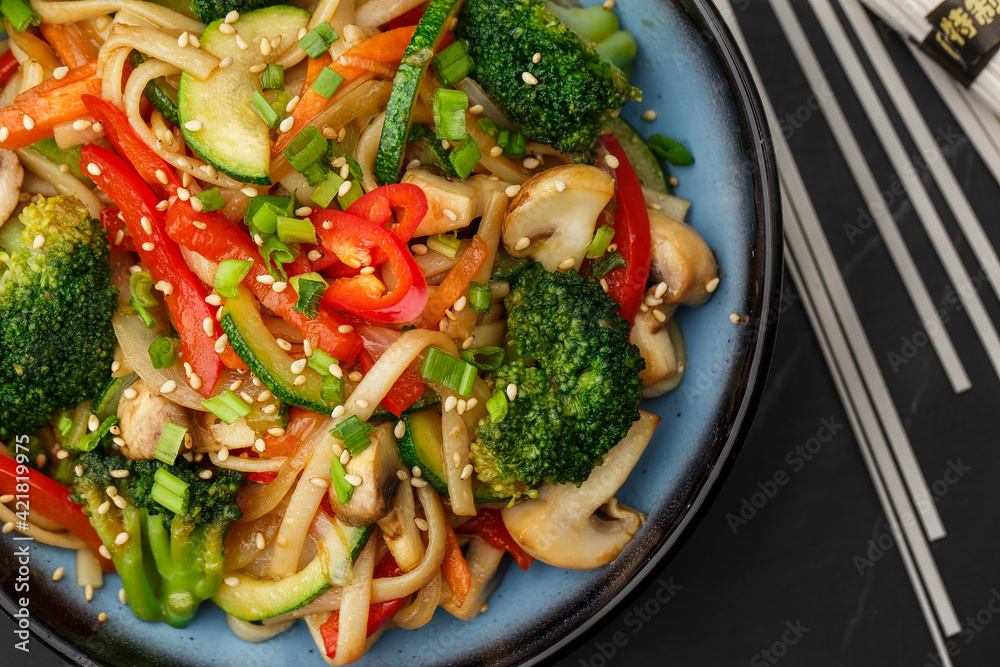 Udon noodles with vegetables: zucchini, broccoli, red bel pepper, mushrooms, carrot, spring onion and sesame seeds. Dish isolated in a blue bowl, close-up on a black marble background. Asian cuisine. Udon noodles with vegetables: zucchini, broccoli, red bel pepper, mushrooms, carrot, spring onion and sesame seeds. Dish isolated in a blue bowl, close-up on a black marble background. Asian cuisine.