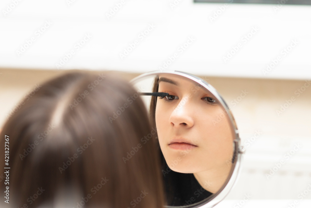 Fototapeta premium Young woman putting on makeup while looking in the mirror paints eyelashes mascara for the eyes