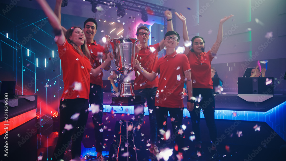 Fototapeta premium Diverse Esport Team Winner of the Video Games Tournament Celebrates Victory Cheering and Holding Trophy in Big Championship Arena. Cyber Gaming Event with Gamers and Fans.
