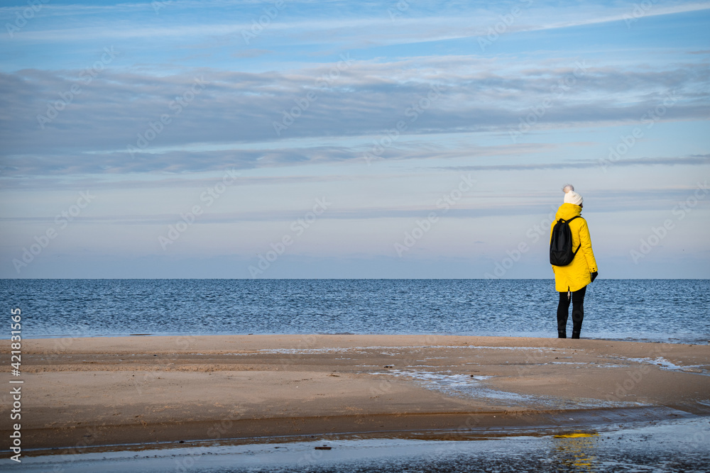 Obraz premium A woman in a yellow winter jacket and with a black backpack walking by the shores of the Baltic Sea near Carnikava, Latvia
