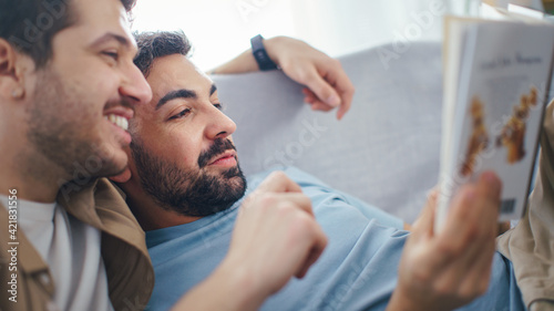 Happy Gay Couple in Love Spending time at Home, Reading Book Together. Boyfriends Lying Together, Embracing Gently, Lovingly. They talk, Have fun. Close-up Portrait.