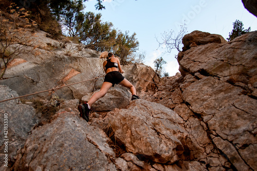 bottom view of athletic young woman who climbing up on rocks using rope.