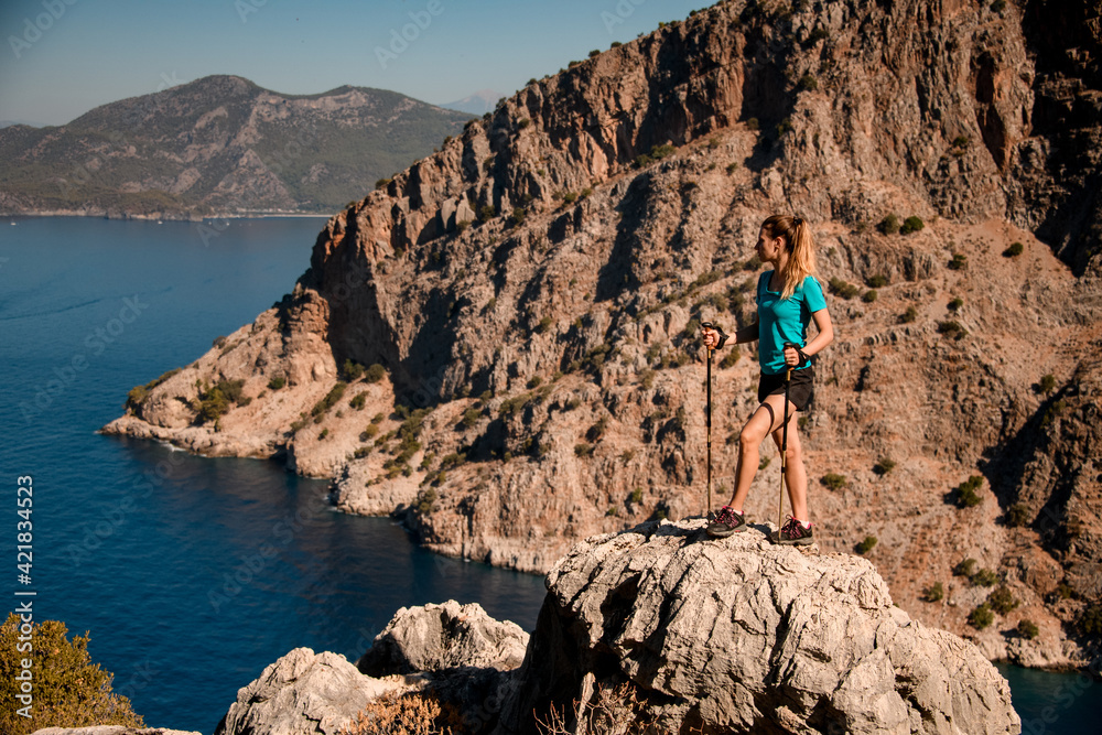 Woman with tracking poles stands on large stone and enjoying view of mountains and sea.