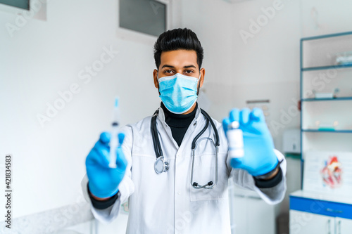 An Indian male doctor in a protective mask and blue glove holds an injection and vaccine in his hands and points to the camera. Modern hospital. Vaccination