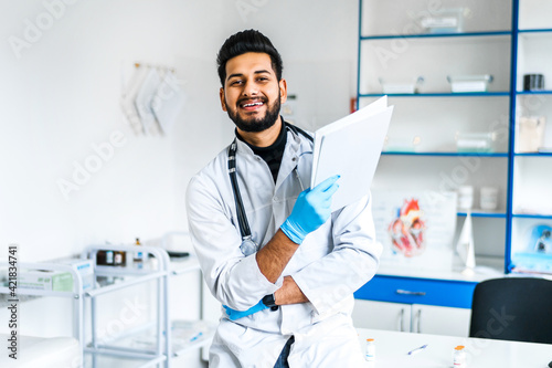 A smiling Indian doctor in the middle of a modern hospital room with a stethoscope and documents in his hands looks into the camera and smiles, an Indian man