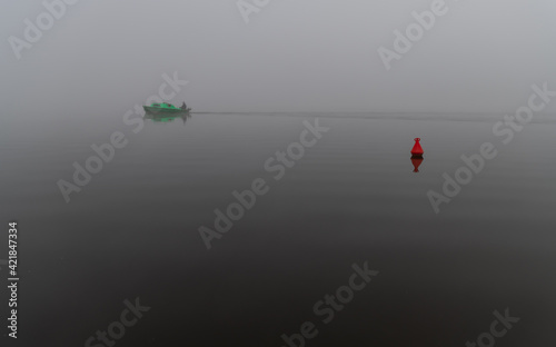See am frühen Morgen im Nebel mit einer Boje und grünem Boot. Ein nebliger Morgen am See.