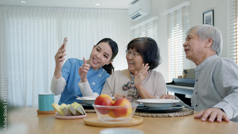 Young senior asia citizen couple and assisted home living nurse care hold mobile phone selfie videocall online at home apartment on dining table in virtual digital telehealth telemedicine concept. Young senior asia citizen couple and assisted home living nurse care hold mobile phone selfie videocall online at home apartment on dining table in virtual digital telehealth telemedicine concept.