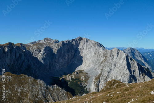 A panoramic view on Hochschwab mountain chains from the pathway leading to Hohe Weichsel. There is a vast pasture on top of a mountain, slowly turning golden. Clear view. Blue sky above. Autumn vibe