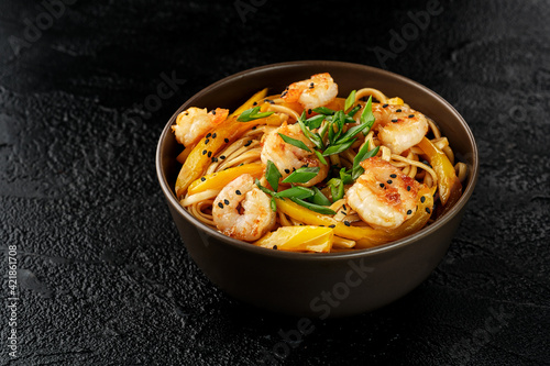 Wok egg noodles with shrimps, yellow pepper and sesame seeds in a dark brown plate on a black background. Asian food. View from above.