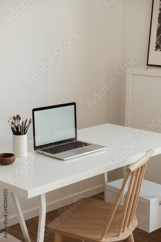Laptop with blank copy space screen on table with stationery on table against white wall. Minimalist home office workspace. Mockup template.