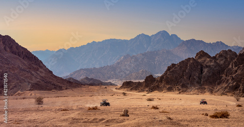 Fototapeta Naklejka Na Ścianę i Meble -  Mountain landscape in a stone desert with silhouettes of high hills at sunset. Extreme adventures on tourist transport in desert, off-road trips and active leisure for tourist in Egypt.