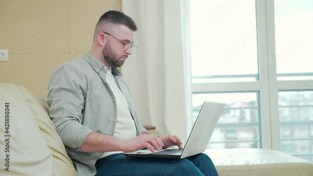 young handsome bearded business man in glasses working with laptop sitting on sofa or couch at home in living room. Male businessman freelancer typing computer online office. Remote work distance young handsome bearded business man in glasses working with laptop sitting on sofa or couch at home in living room. Male businessman freelancer typing computer online office. Remote work distance