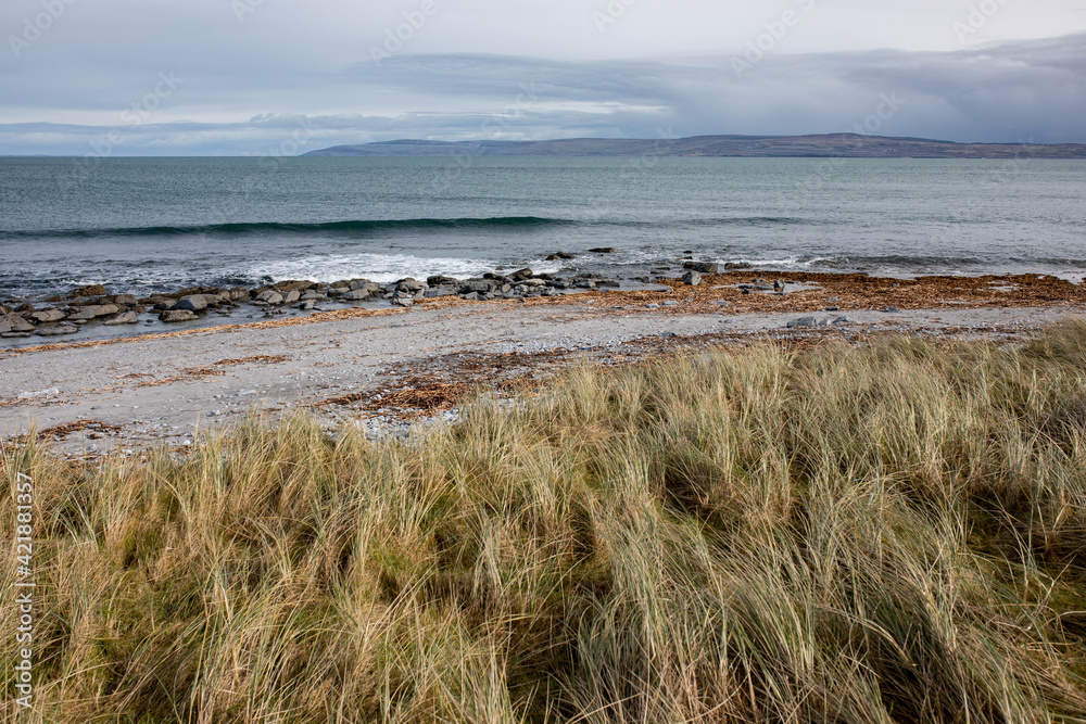 Landscape with grey pebble beach, dunes, beige dry grass, blue ocean and sunny, blue sky on Inisheer island, the smallest of Aran islands. Landscape with grey pebble beach, dunes, beige dry grass, blue ocean and sunny, blue sky on Inisheer island, the smallest of Aran islands.
