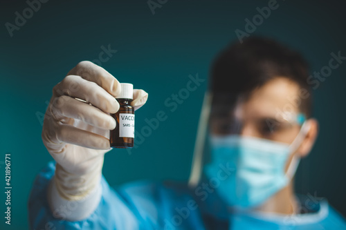 doctor in a protective shield, veil, and sterile gloved suit holds a single weapon against the Coronavirus pandemic. A scientist holds an ampoule against a Covid-19 and points to the camera