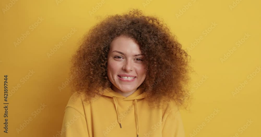 Pretty optimistic millennial European woman with natural curly bushy hair smiles pleasantly expresses happy emotions wears sweatshirt isolated over yellow background. Sincere emotions concept Pretty optimistic millennial European woman with natural curly bushy hair smiles pleasantly expresses happy emotions wears sweatshirt isolated over yellow background. Sincere emotions concept