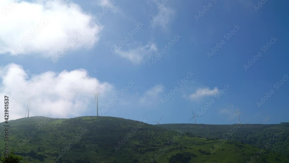 Wind generators revolving on hill with mountains and clouds with blue sky in background. Environment conservation concept. Alternative means of energy concept 4K footage.