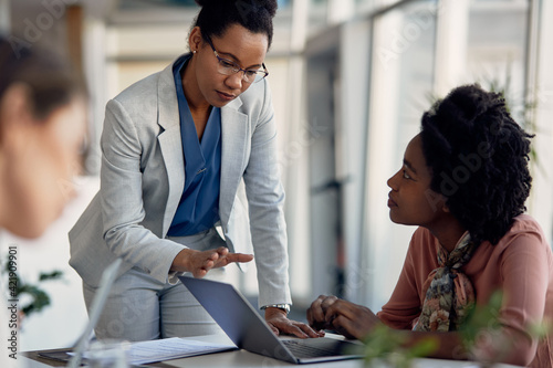 African American businesswoman coaches younger colleague who works on computer at corporate office.