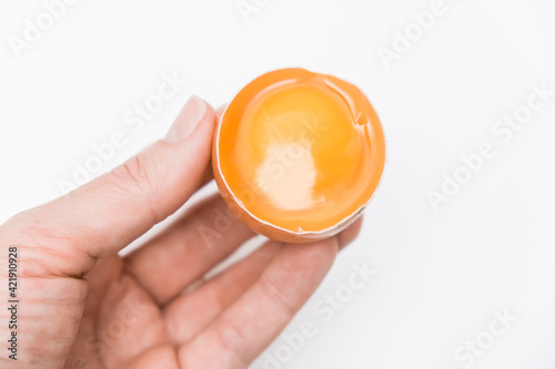 A hand holding half broken egg with yellow yolk in a shell against white background close up
