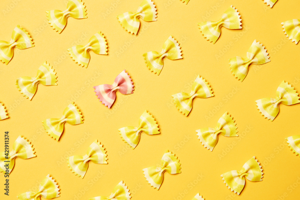 Obraz premium Pink bow tie butterfly farfalle shape pasta one alone single piece stands out of yellow pieces diagonally oriented pattern top view isolated on the bright solid yellow fond background