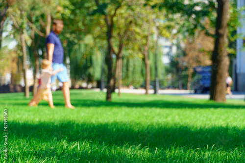 people walking and children playing in a city park on a summer day, green lawns with grass and trees, paths and benches, bright sunlight and shadows