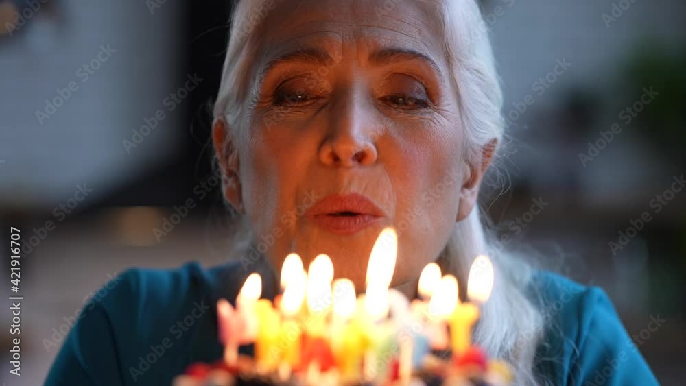 Close-up of attractive cheerful elderly woman with white hair blowing candles on colored Happy birthday lettering during anniversary celebration. Portrait of happy gray-haired female during holiday