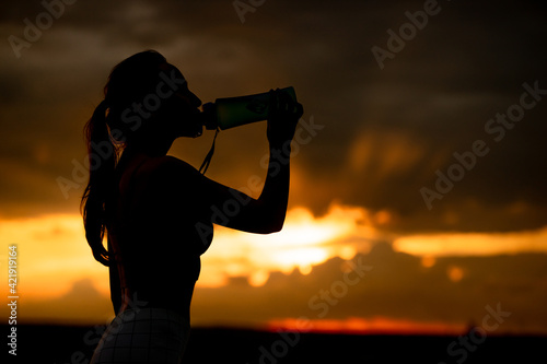 Silhouette of girl in sportswear drinks water from a bottle at sunset